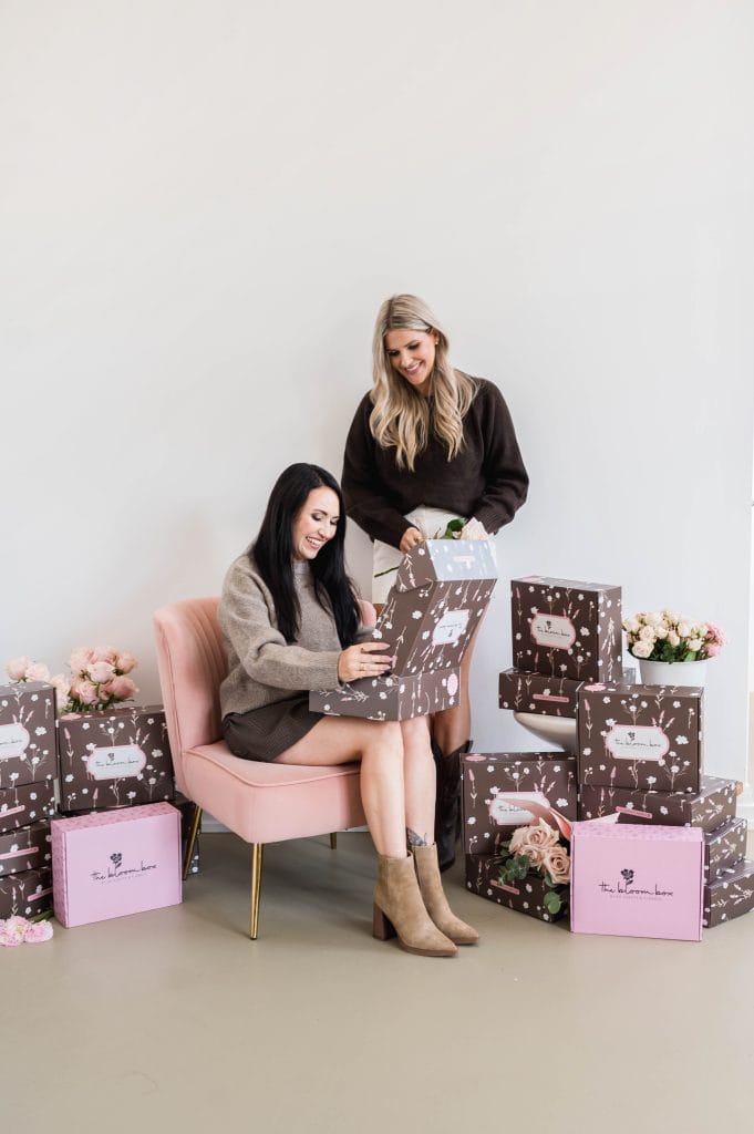 Two women with floral gift boxes and bouquets in a bright studio.