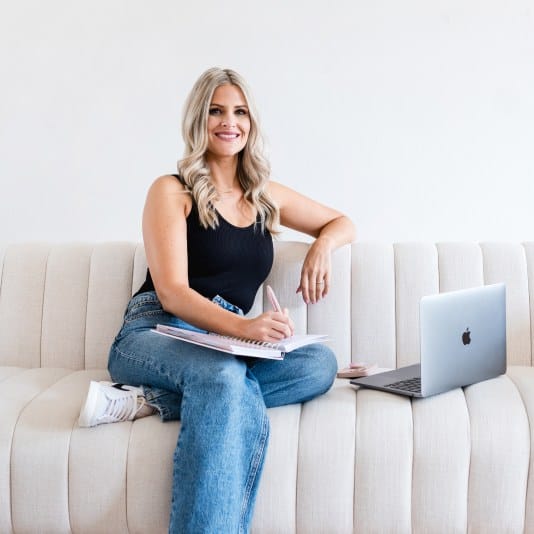 Young woman sitting on sofa with laptop and notebook, smiling at camera.