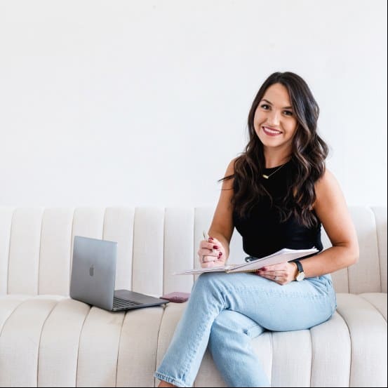 Young woman with dark hair smiling, sitting on a white sofa with a laptop and notebook.