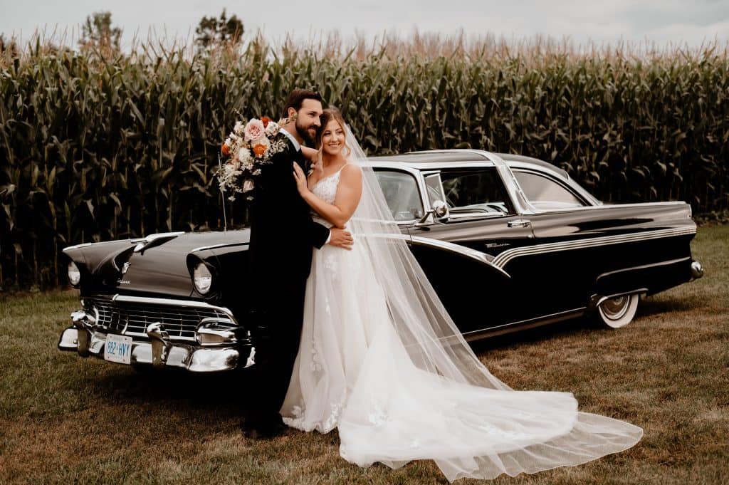 Bride and groom with vintage black car and floral bouquet at wedding.