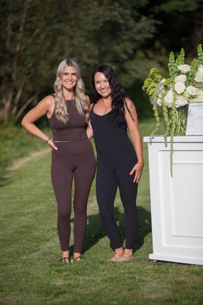Two women in black outfits at outdoor floral event with white floral arrangements.