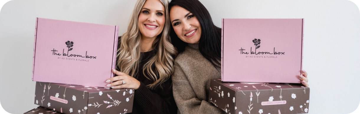 Two women holding pink floral gift boxes from XO Flowers, smiling.