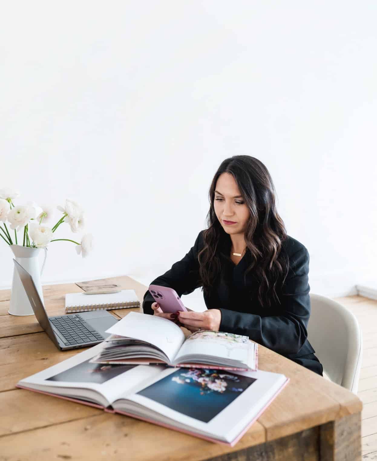Businesswoman using smartphone with floral decor and open photo albums on desk.