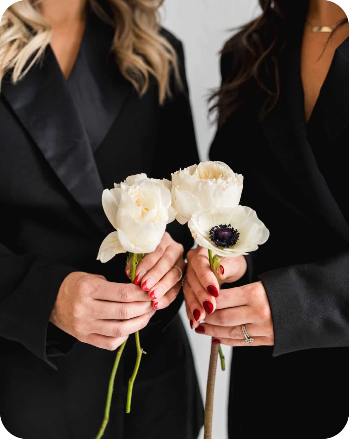Two women holding white and purple floral bouquets in black attire.