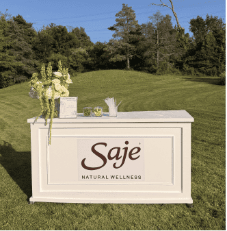 Outdoor event bar with floral arrangements and Saje Natural Wellness branding.