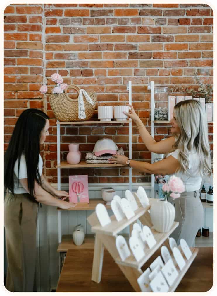 Two women arranging pink and white flowers in a floral shop with brick wall background.